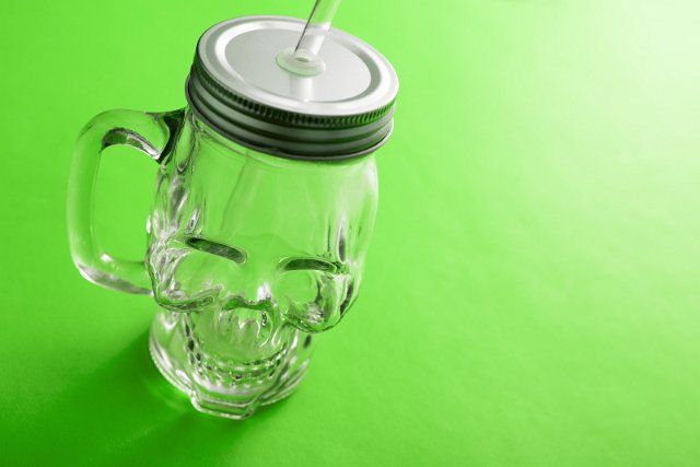 Empty smiling Halloween theme skull glass mug covered by lid and straw over green background with copy space