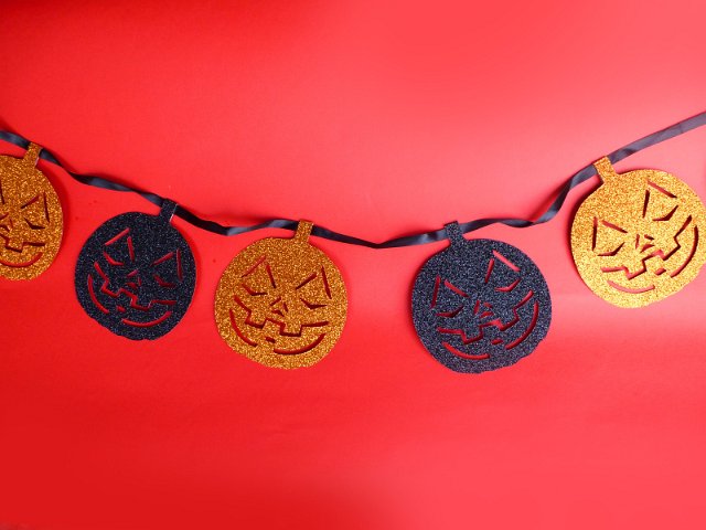 Halloween decoration of evil smiling pumpkin faces in orange and blue glitter hanging on a string over festive red background with copy space